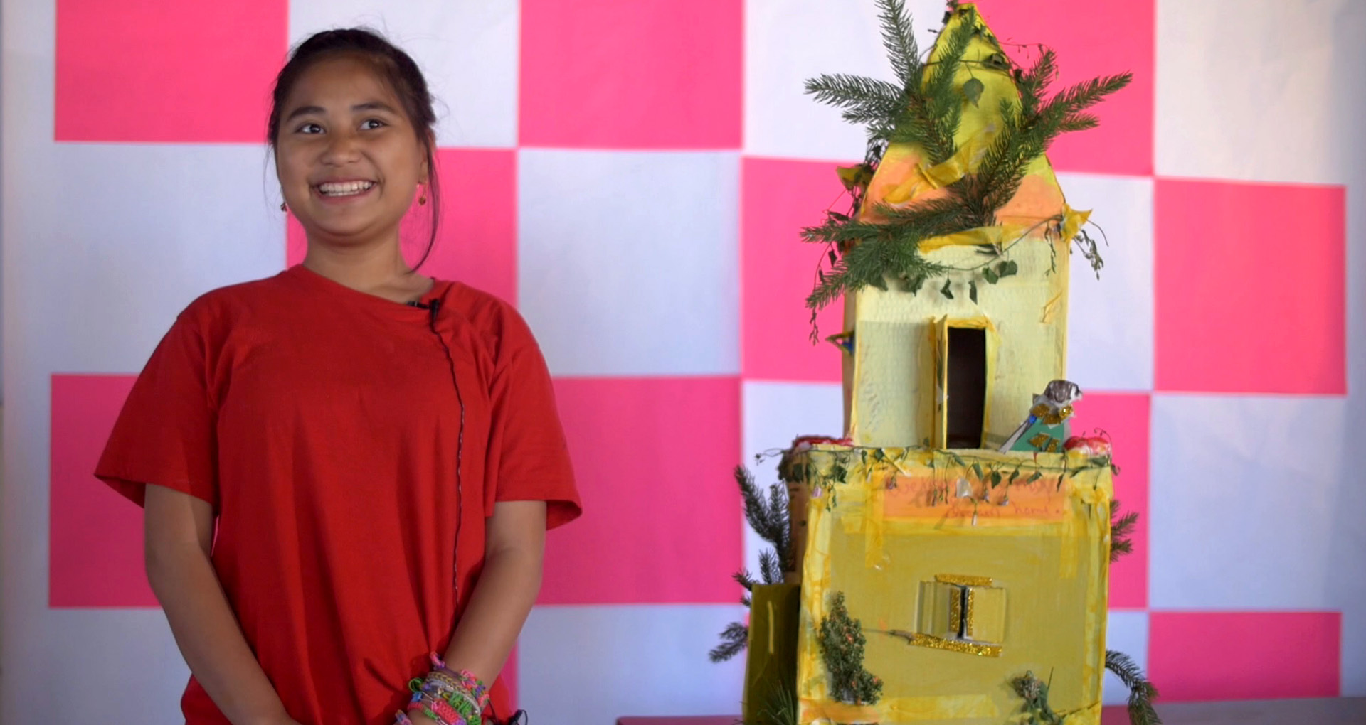 little girl in red tshirt in front of a pink and white background, in front of a cardboard castle painted yellow